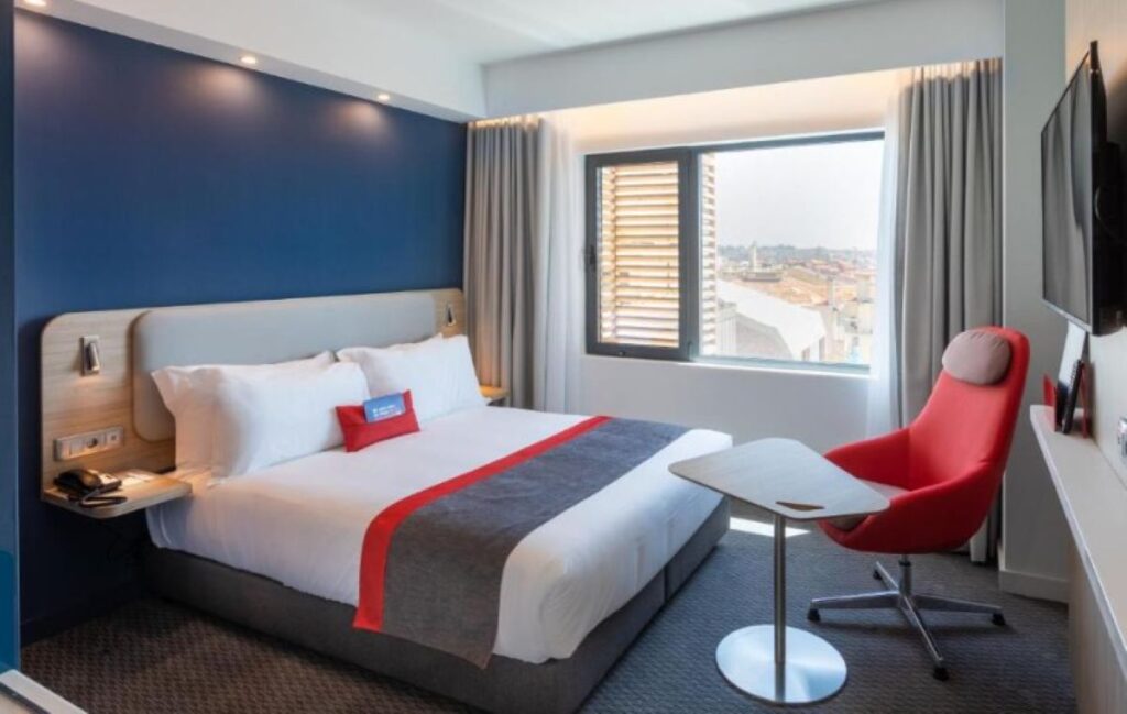 Modern hotel bedroom with navy blue accent wall, red designer chair, and wooden shutters framing city views