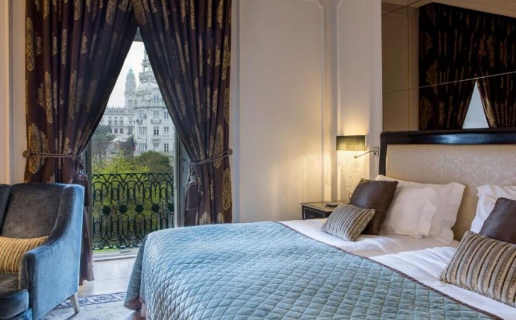 Elegant hotel bedroom with ornate curtains, wrought iron balcony, and views of Porto's historic architecture through French doors