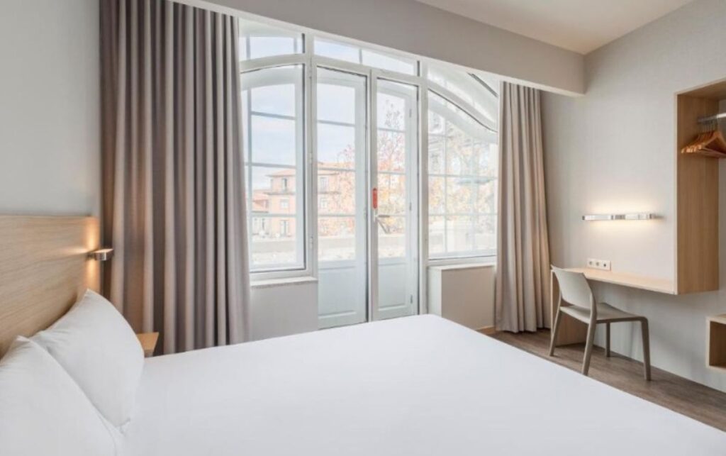 Minimalist hotel room with white bedding, wooden desk area, and floor-to-ceiling windows opening to balcony