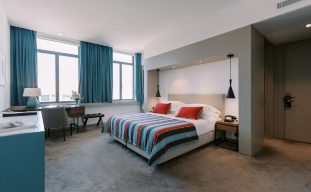 Spacious hotel suite with teal curtains, striped bedding, pendant lighting, and dedicated workspace by large windows
