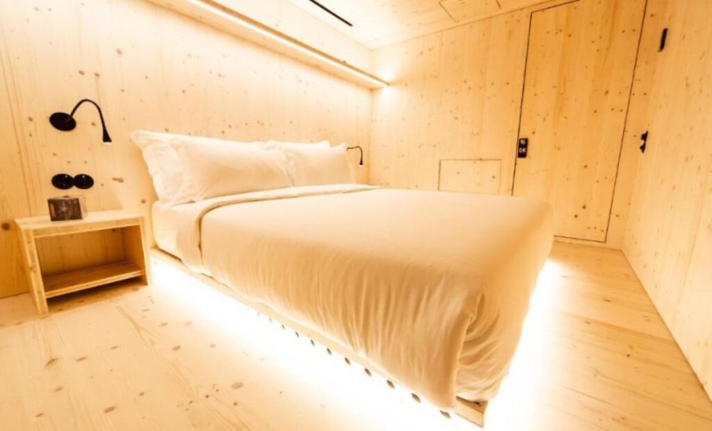 Compact wooden pod-style hotel room with built-in storage, warm lighting, and natural pine panelling throughout