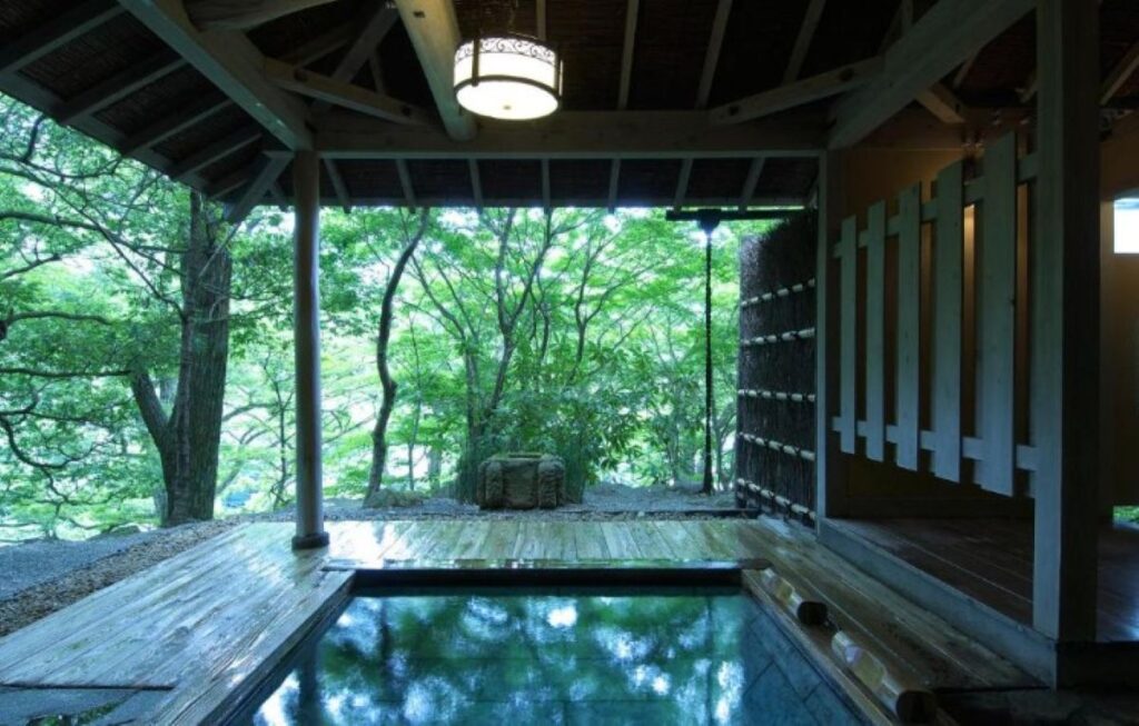 Semi-outdoor wooden onsen bath open to forest surroundings beneath traditional pavilion