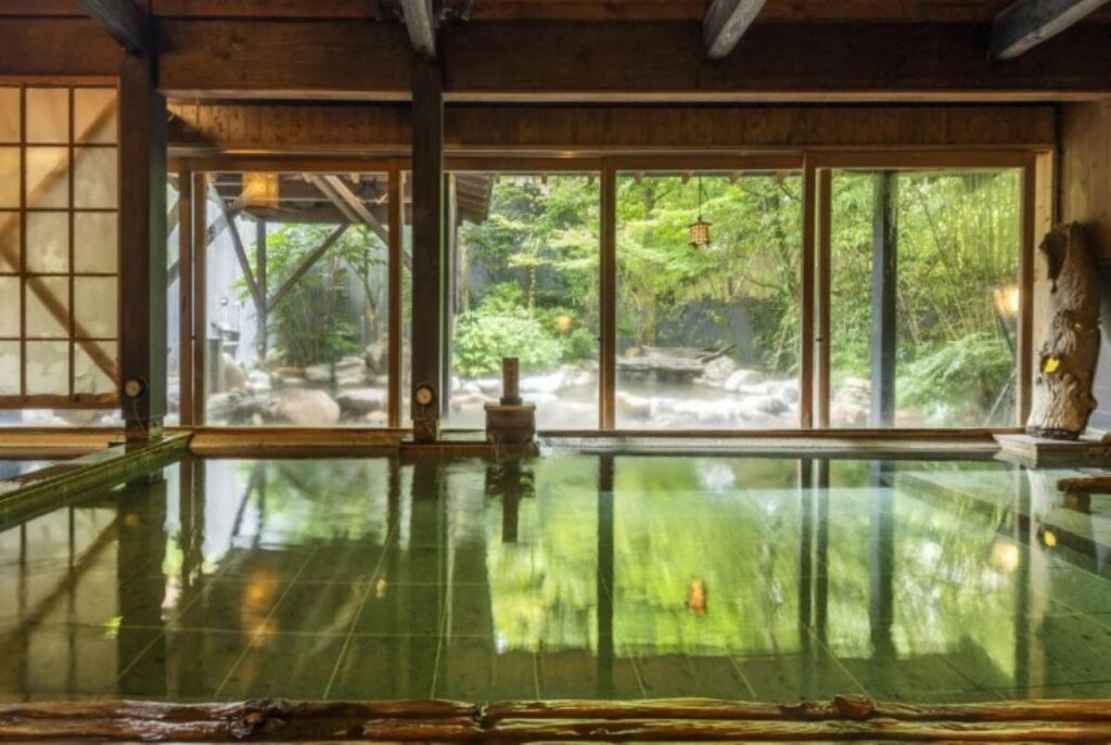 Indoor onsen bath with wooden beams and floor-to-ceiling windows overlooking Japanese garden