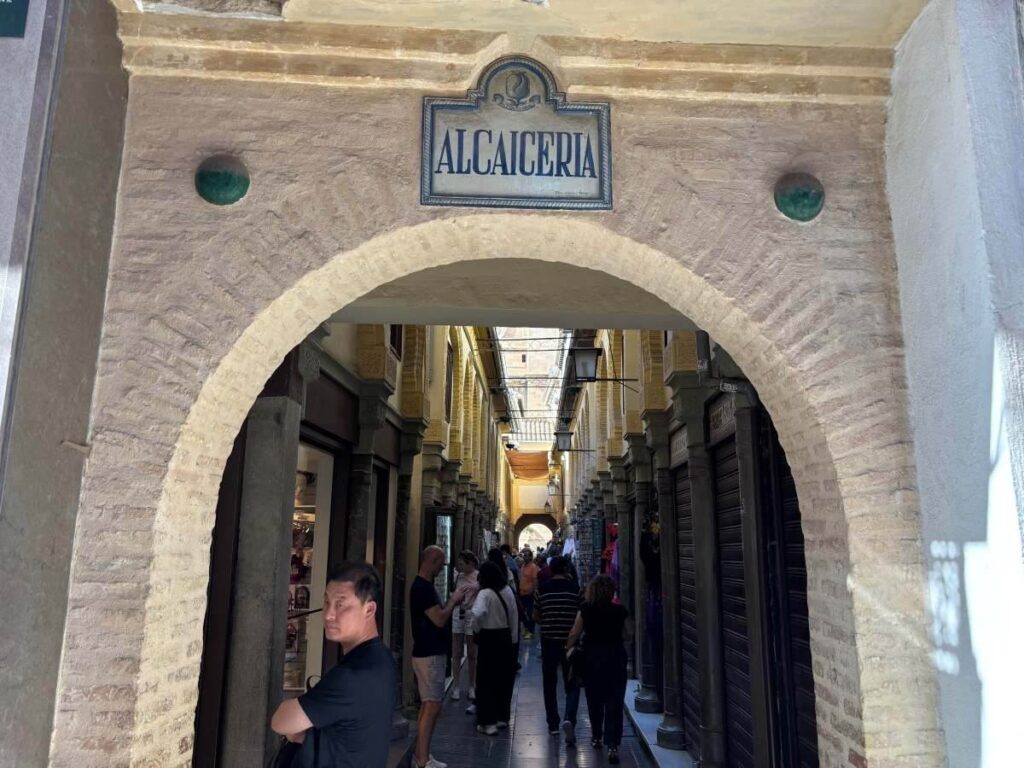 Entrance arch to the Alcaicería market in Granada leading into a narrow arcade of shops and people.