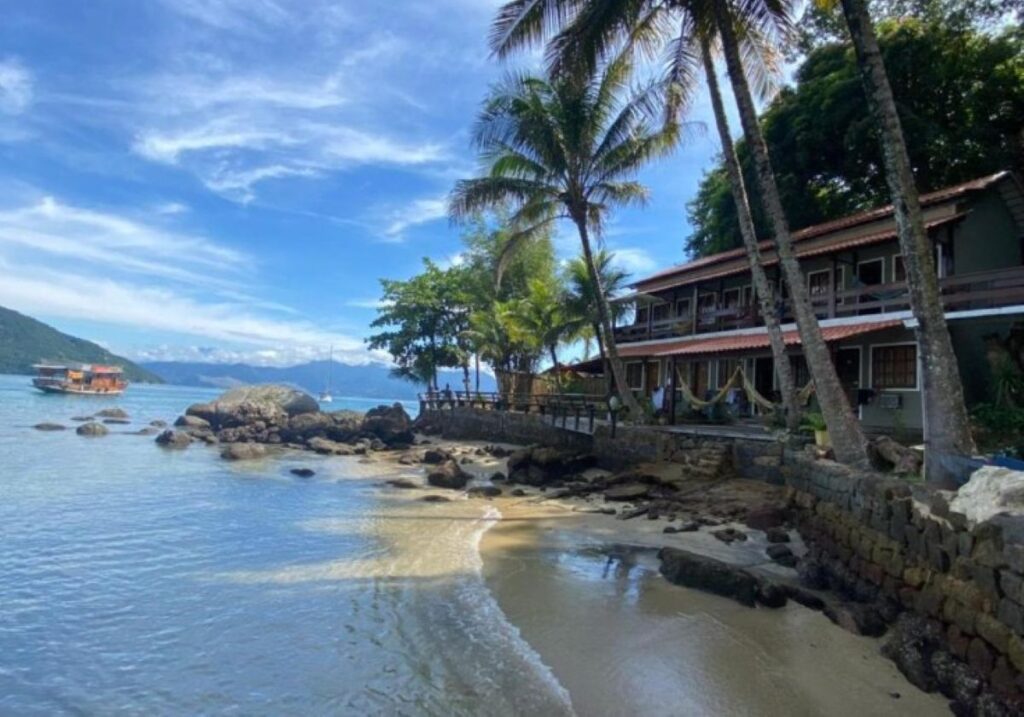Beachfront pousada building with palm trees, rocky shoreline and mountain backdrop