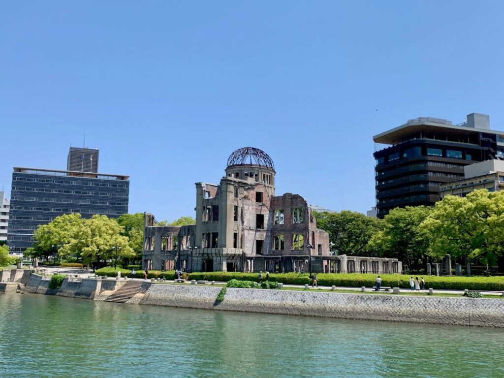 Hiroshima’s A Bomb Dome beside the river, the ruined brick building standing among green trees and modern offices on a sunny day.