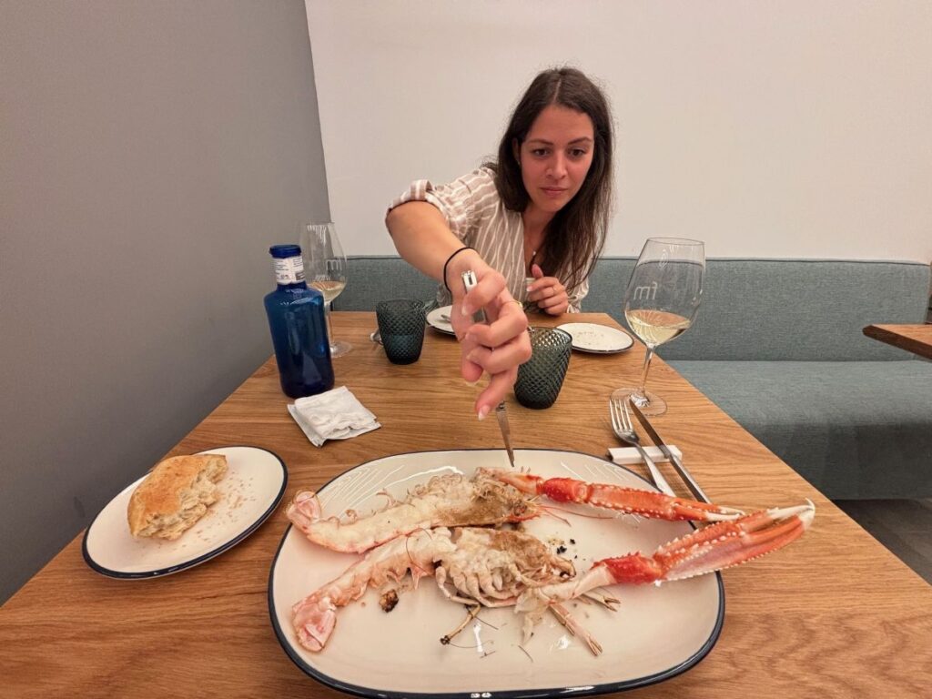Woman cutting into grilled langoustine at a restaurant table with wine glasses and bread.