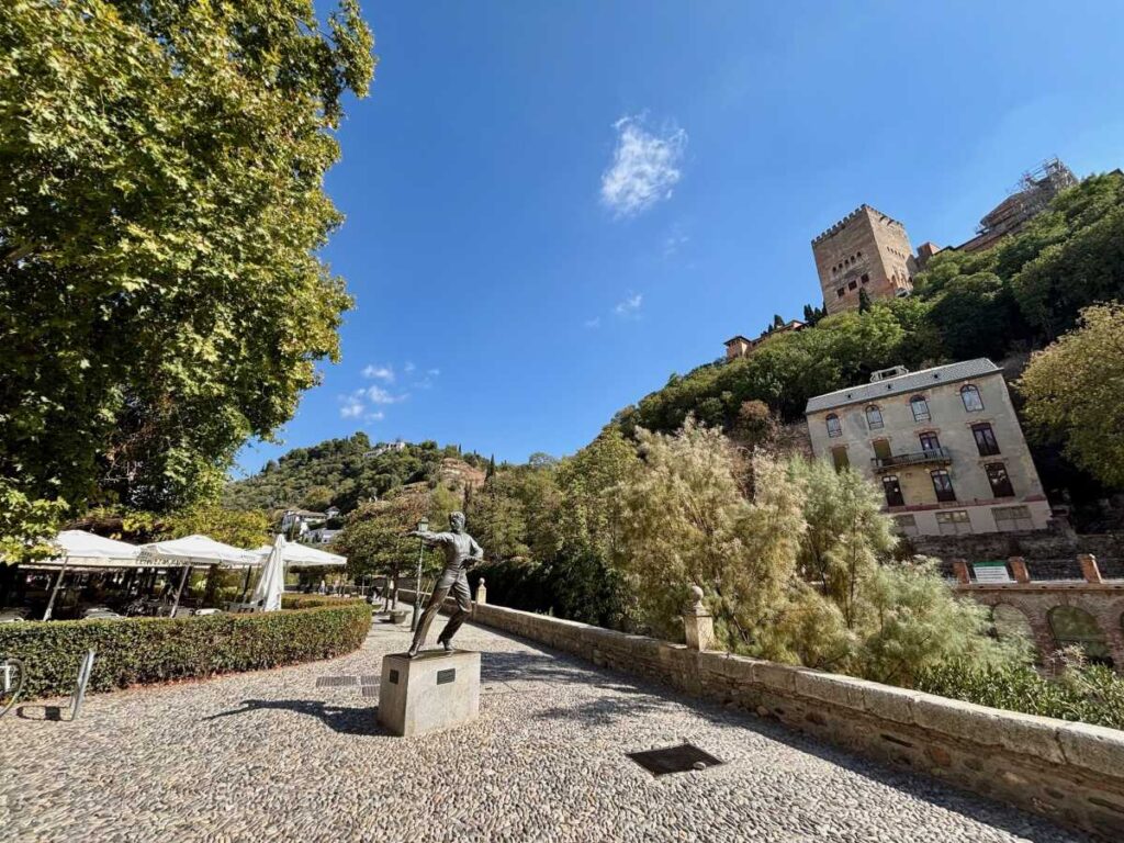 Statue on the riverside paseo below the Alhambra, with cafés, trees and old houses on the hillside.