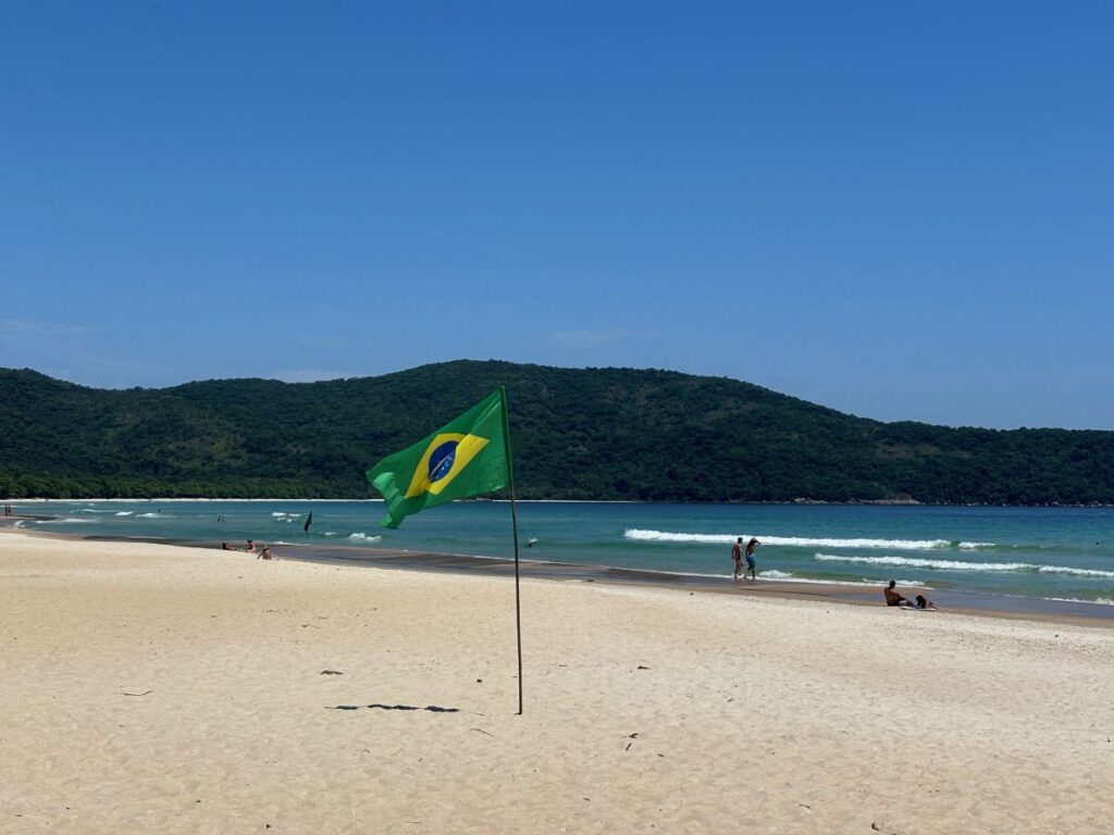 Brazilian flag planted in the sand on a wide Ilha Grande beach with calm waves and forested hills behind