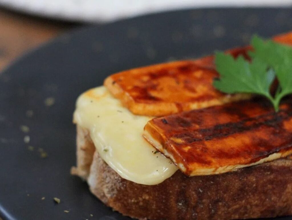 Open sandwich topped with melted cheese and grilled halloumi on dark slate