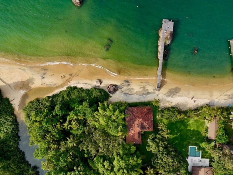 Aerial view of a quiet Ilha Grande beach with a wooden jetty, shallow green water, and dense tropical forest behind the sand