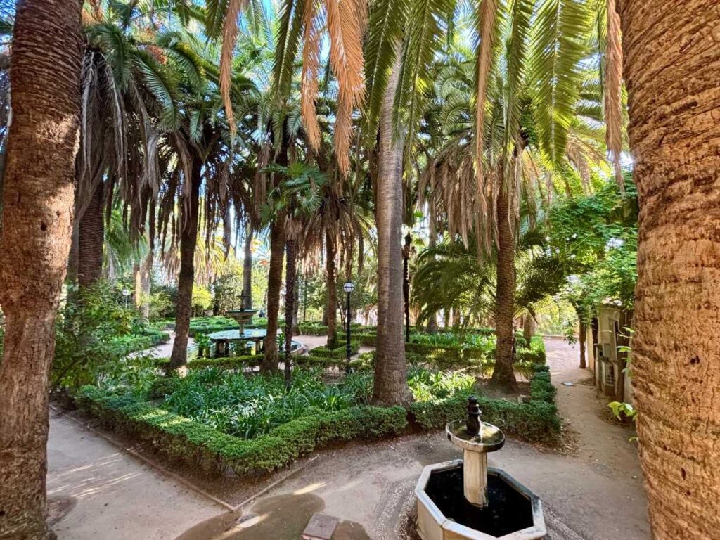 Shaded palm garden in Granada with neat flower beds, winding paths and two small stone fountains.