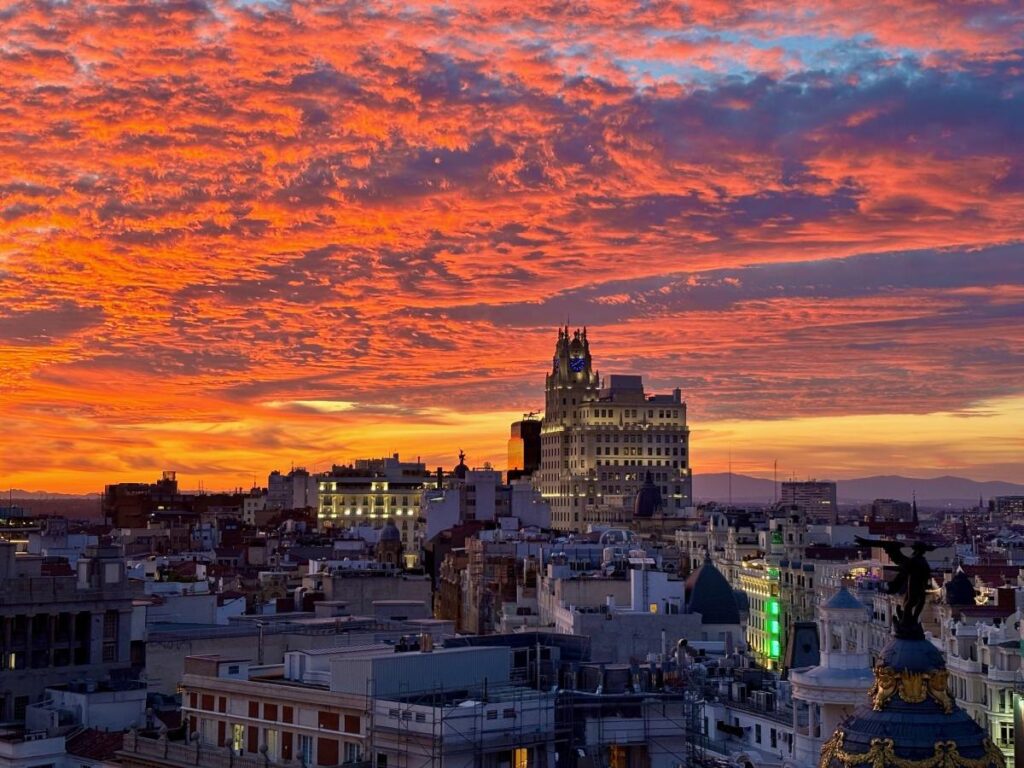 Fiery Madrid sunset over rooftops and lit streets, with the Metropolis dome in the foreground and the skyline glowing under orange clouds.