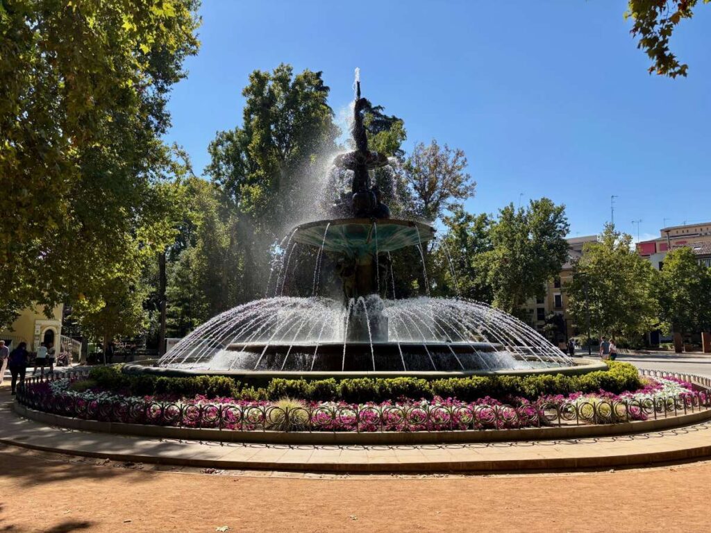 Large circular fountain in a Granada square, ringed with flowers and trees under a bright blue sky.