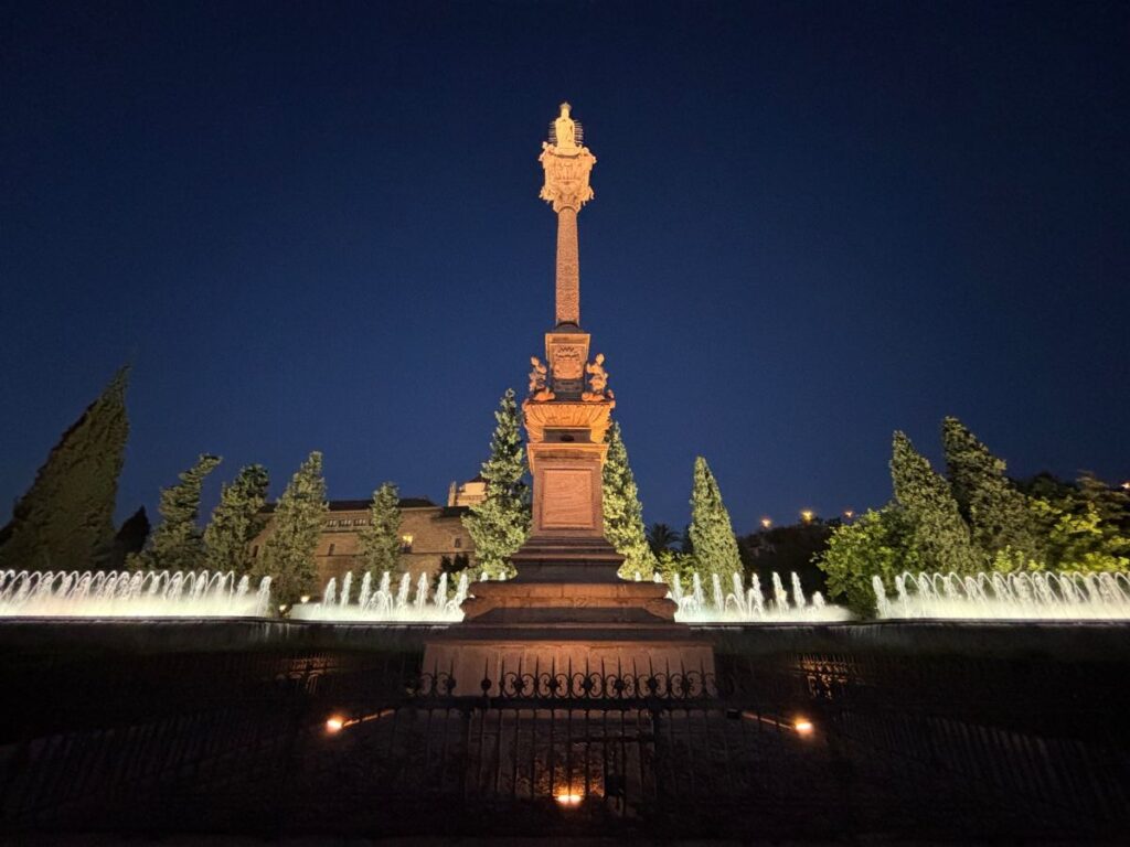 Floodlit stone column and statue surrounded by arcing fountains in a park at night in Granada.