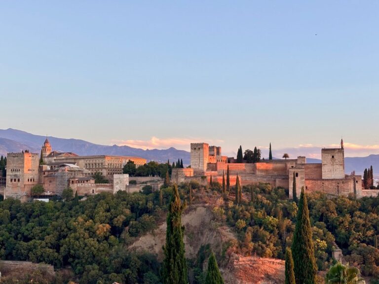 Sunset view of the Alhambra palace and fortress above Granada, with cypress trees and Sierra Nevada mountains behind.