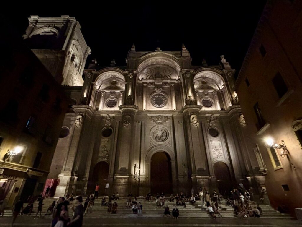 20. Night view of Granada Cathedral’s illuminated façade with people gathered on the wide front steps.