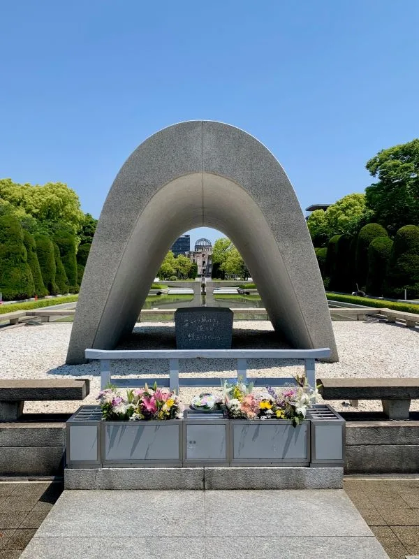 Hiroshima Peace Memorial Cenotaph with fresh flower tributes, framing the distant A Bomb Dome under a clear blue sky.