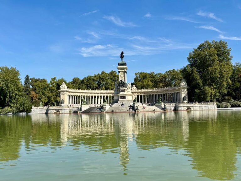 El Retiro Park lake in Madrid with the Monument to Alfonso XII reflected in the water, framed by trees on a bright day.