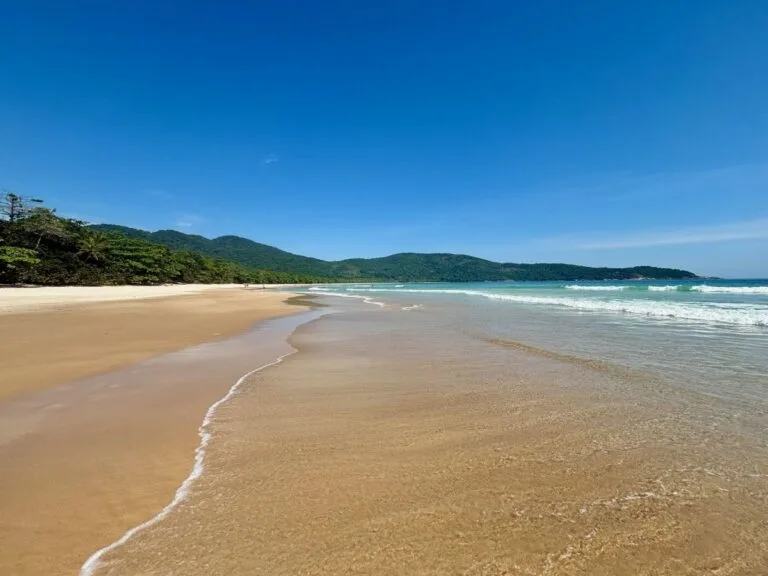 Empty Ilha Grande beach with smooth sand, clear water, and low forested hills under a bright blue sky