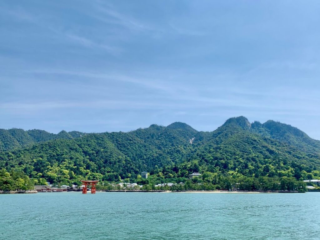 View from the ferry to Miyajima, with the red Itsukushima Shrine torii gate standing in the sea below forested mountains and a hazy blue sky.