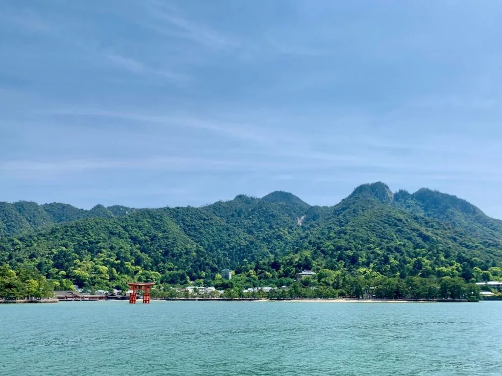 View from the ferry to Miyajima, with the red Itsukushima Shrine torii gate standing in the sea below forested mountains and a hazy blue sky.