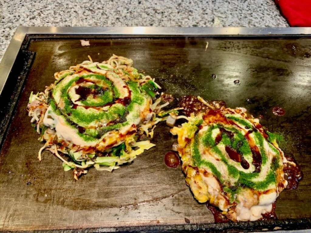 Okonomiyaki cooking on teppan grill with green aonori, mayonnaise and brown sauce drizzle