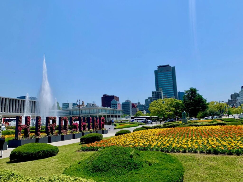 Colourful flower beds, fountains and a large HIROSHIMA sign in Peace Memorial Park on a bright spring day, modern city skyline in the background.