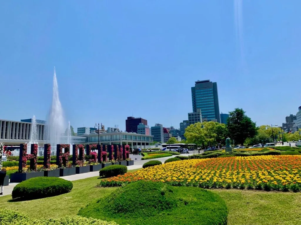 Colourful flower beds, fountains and a large HIROSHIMA sign in Peace Memorial Park on a bright spring day, modern city skyline in the background.