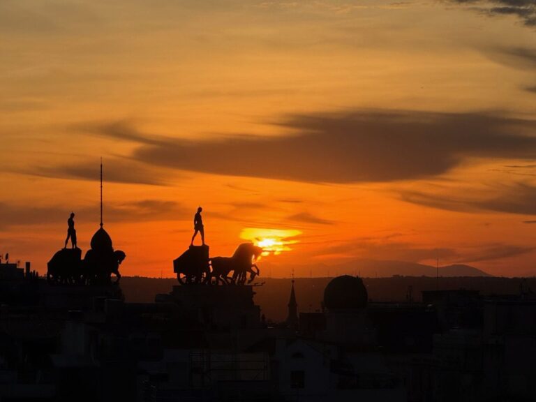 Silhouette of the Metropolis building quadriga in Madrid at sunset, statues outlined against an orange sky and distant hills.