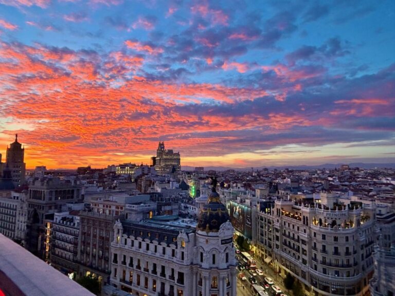 Wide Madrid skyline at dusk, orange and blue clouds stretching over rooftops and bright avenues, photographed from a rooftop viewpoint.