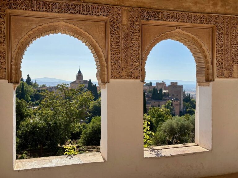 View of the Alhambra and church framed by two ornate Moorish arches, with green trees in the foreground.