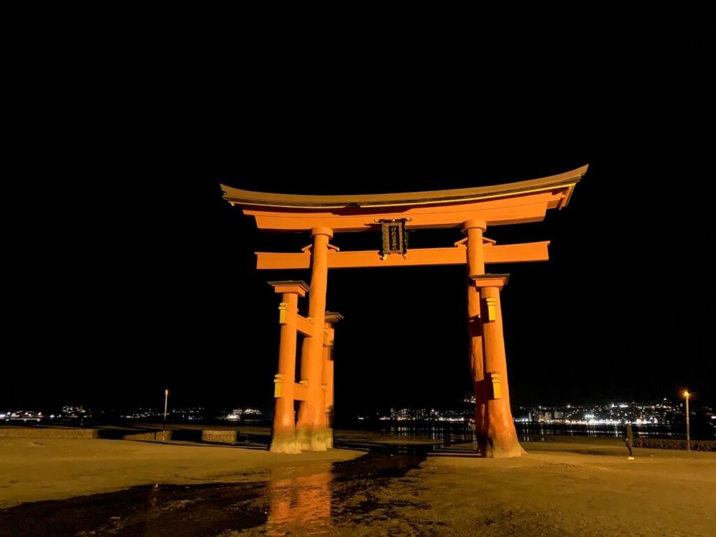 Illuminated Itsukushima Shrine torii gate at night, reflections in wet sand with harbour lights across the water, Miyajima, Japan.