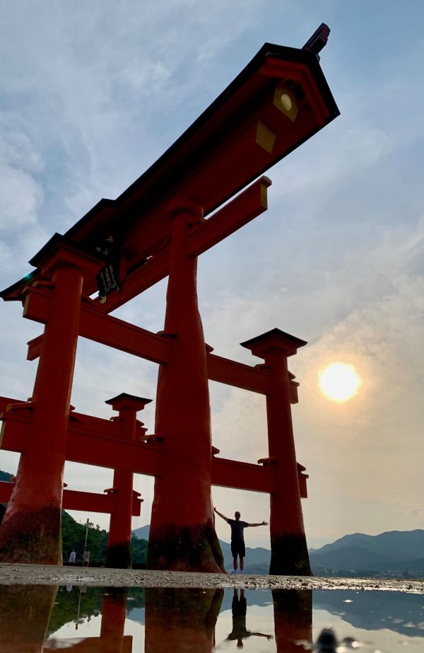 Low-angle view of the giant Miyajima torii gate at sunset, a person beneath it and a puddle reflection on the sand.