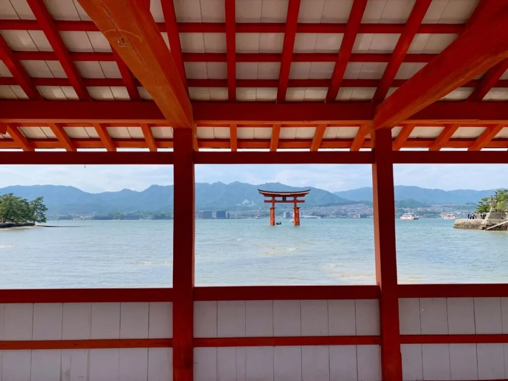 Offshore torii gate framed by red shrine beams, calm sea with mountains and ferries in the distance, Miyajima.