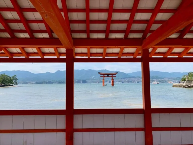 Offshore torii gate framed by red shrine beams, calm sea with mountains and ferries in the distance, Miyajima.