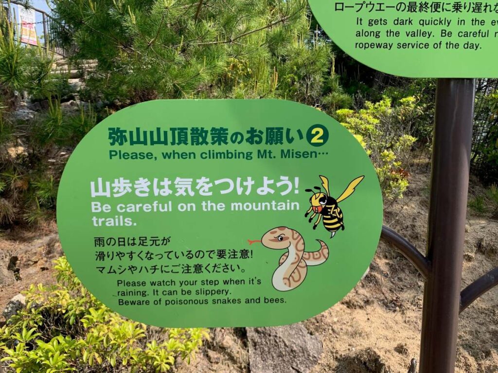 Mt Misen trail warning sign in Japanese and English, with cartoon bee and snake icons and safety notes.