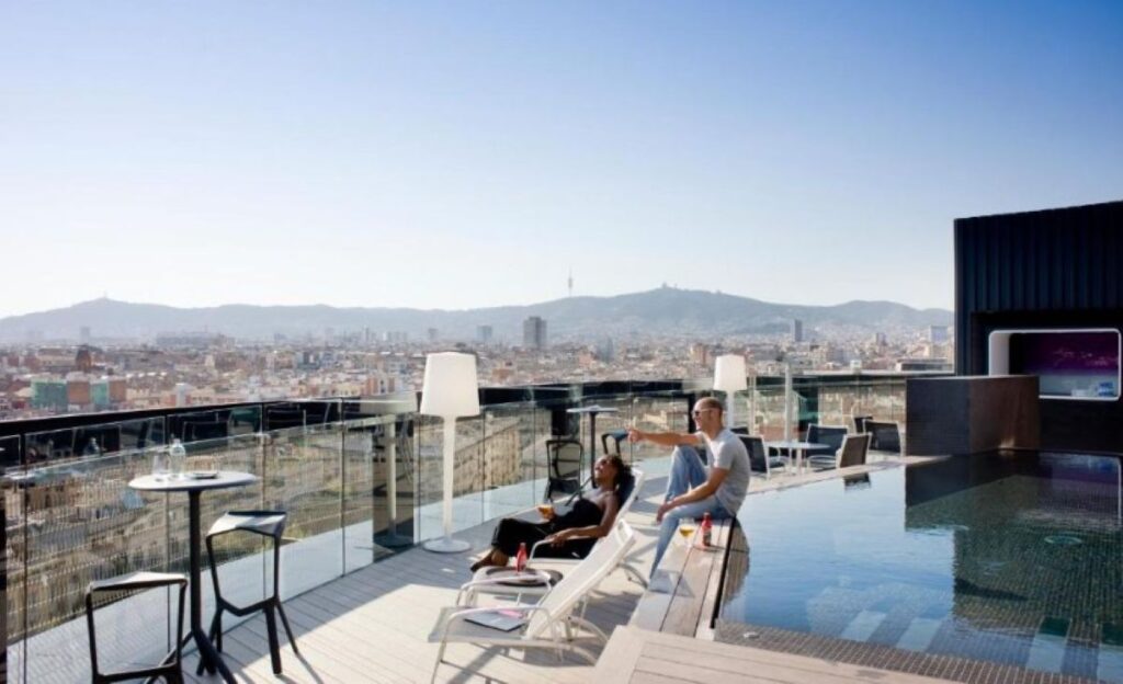Couple relaxing by a rooftop infinity pool on sun loungers with floor lamp and vast city and mountain panorama beyond