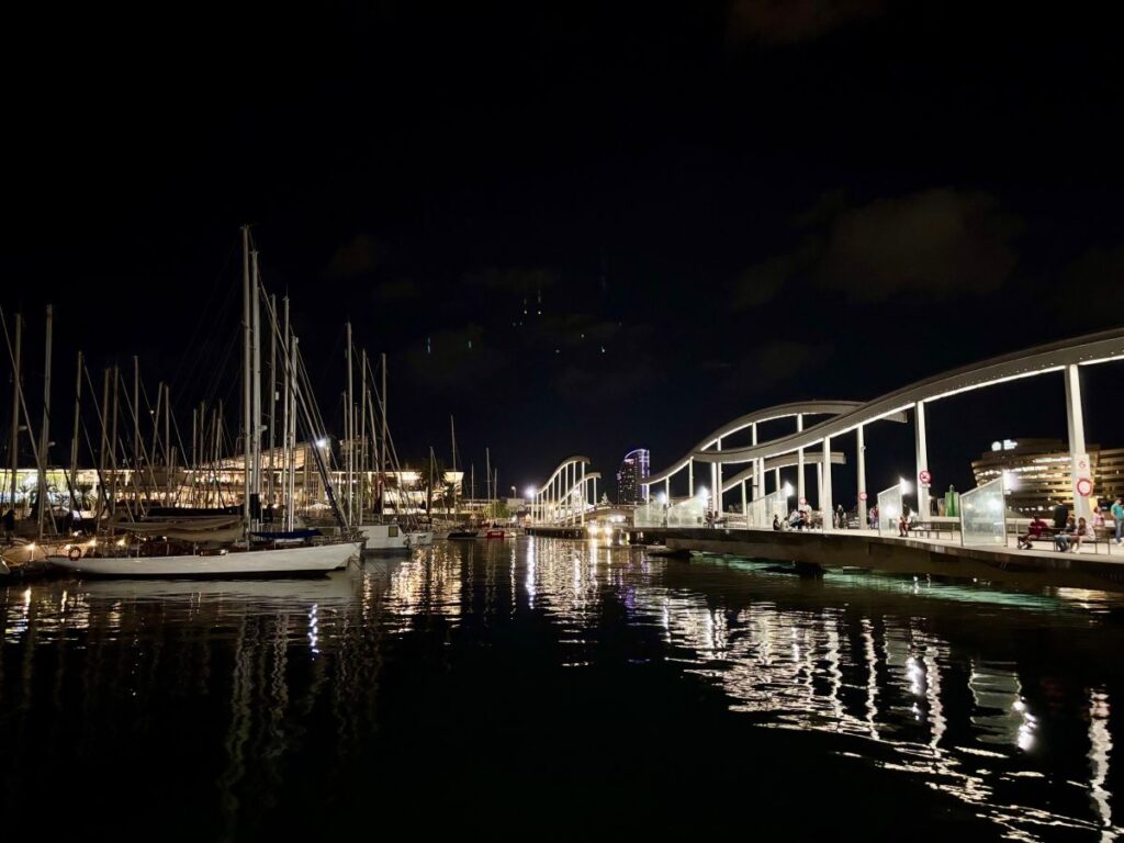 Barcelona marina at night with sailboat masts and a lit pedestrian bridge, reflections shimmering across the water.