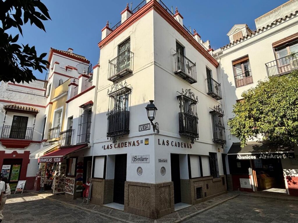 Whitewashed corner building with wrought-iron balconies and shopfronts, sign reading Las Cadenas on a quiet cobbled plaza.