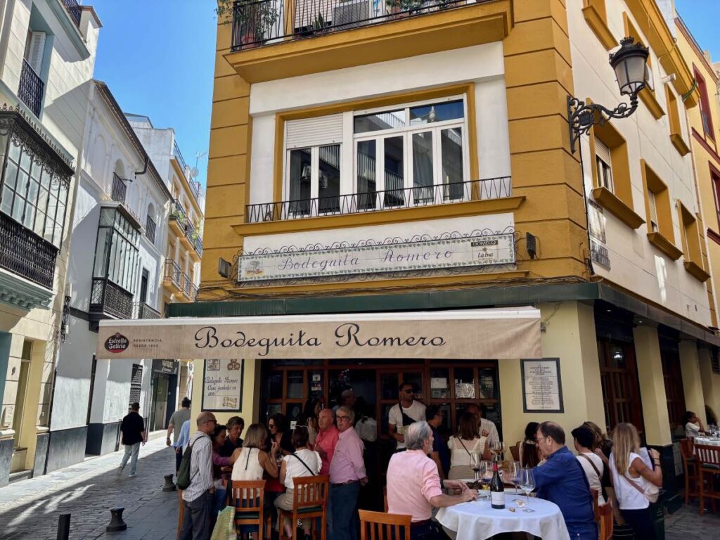 Busy lunchtime scene outside Bodeguita Romero, diners at small tables on a narrow street beneath the café awning and sign.