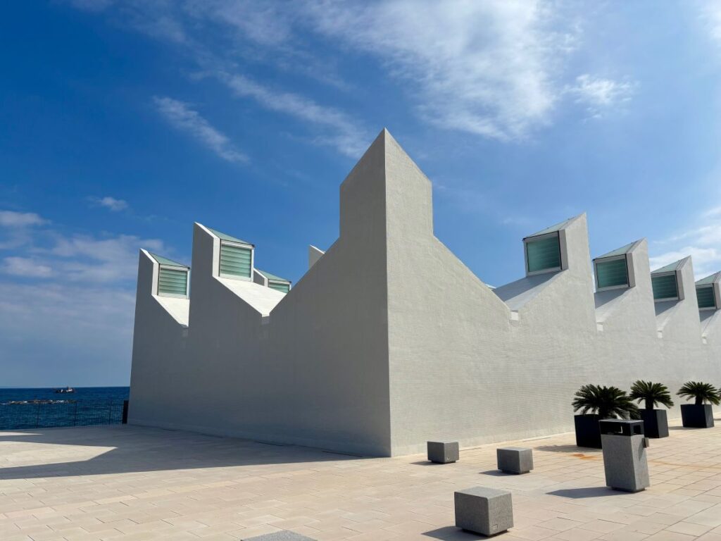 Angular white waterfront building with stepped roofline and skylights, set against blue sky and the Mediterranean.