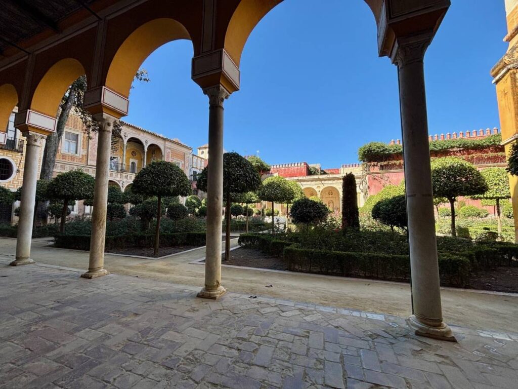 Shaded courtyard at Casa de Pilatos, viewed from under arches, with manicured trees, flowerbeds and sunlit walls beyond.