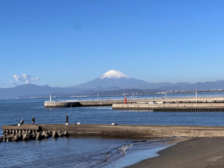Mount Fuji rising beyond Sagami Bay, seen from a seaside breakwater with fishermen and calm blue water.