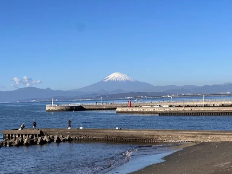 Mount Fuji rising beyond Sagami Bay, seen from a seaside breakwater with fishermen and calm blue water.