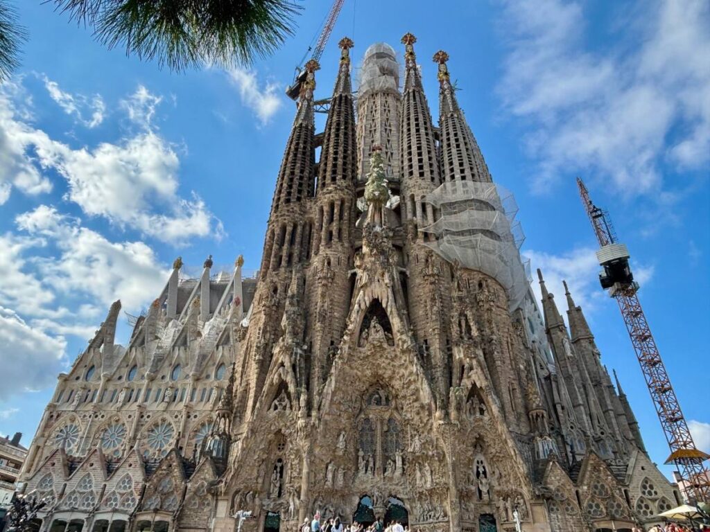 Close-up of the Sagrada Família façade and towers, with cranes and scaffolding against scattered clouds.