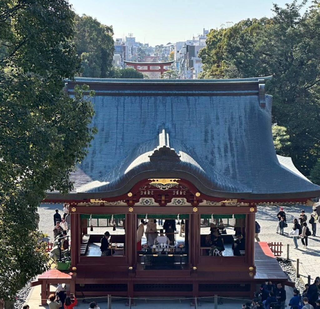 View over a shrine pavilion roof towards a distant torii gate and town streets beyond.