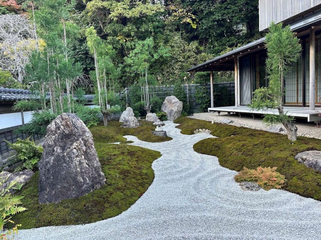Zen rock garden with raked gravel, moss mounds, stones and a small lantern beside a traditional veranda.