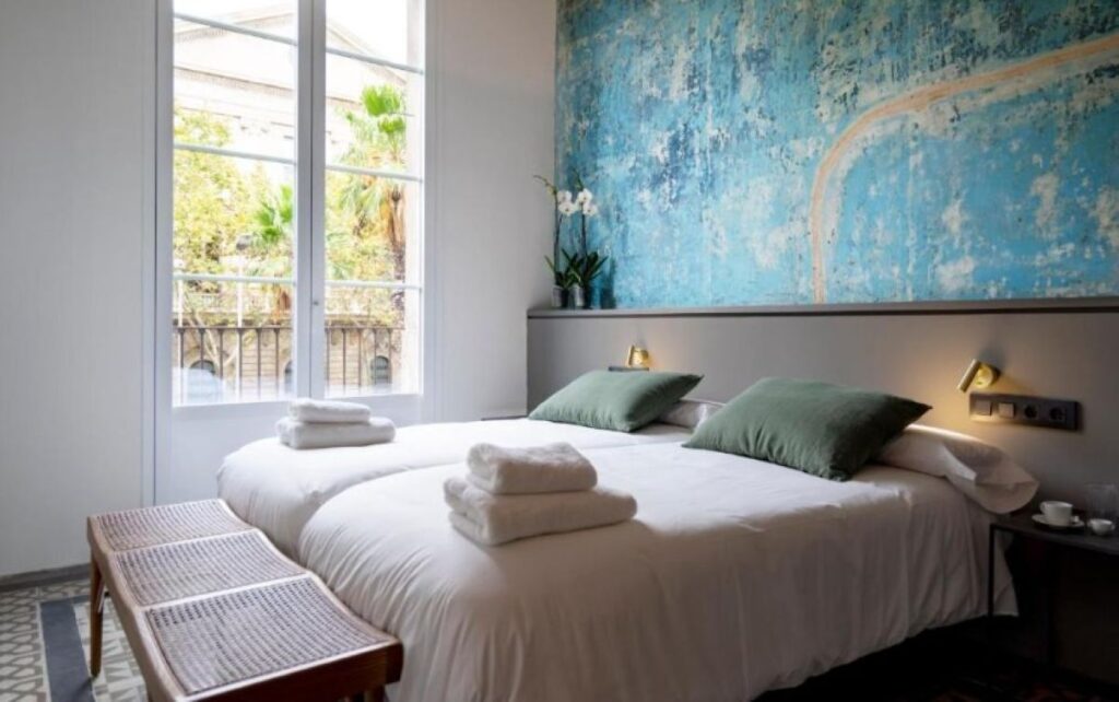 Bright double room with large distressed blue feature wall, sage green cushions, rattan bench and garden balcony