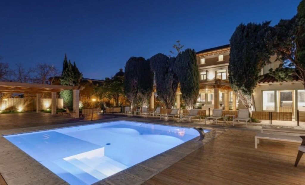 Illuminated outdoor pool at dusk surrounded by cypress trees, timber decking, sun loungers and a lit stone villa